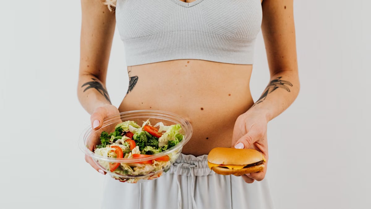 A woman in a sports bra holding a salad and cheeseburger, representing healthy vs junk food choices.